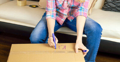 Woman in casual clothes packing a cardboard box with household items in preparation for moving
