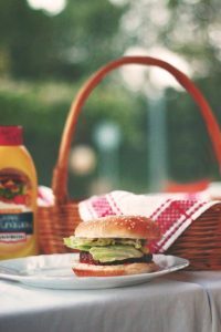 Picnic table set with food, drinks, and a colorful towel on a backyard deck with a bottle of sunscreen and a sandwich