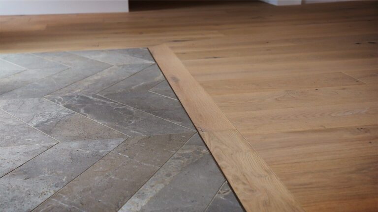 Close-up of a flooring transition between herringbone-pattern stone tiles and natural oak hardwood in an open-concept space.