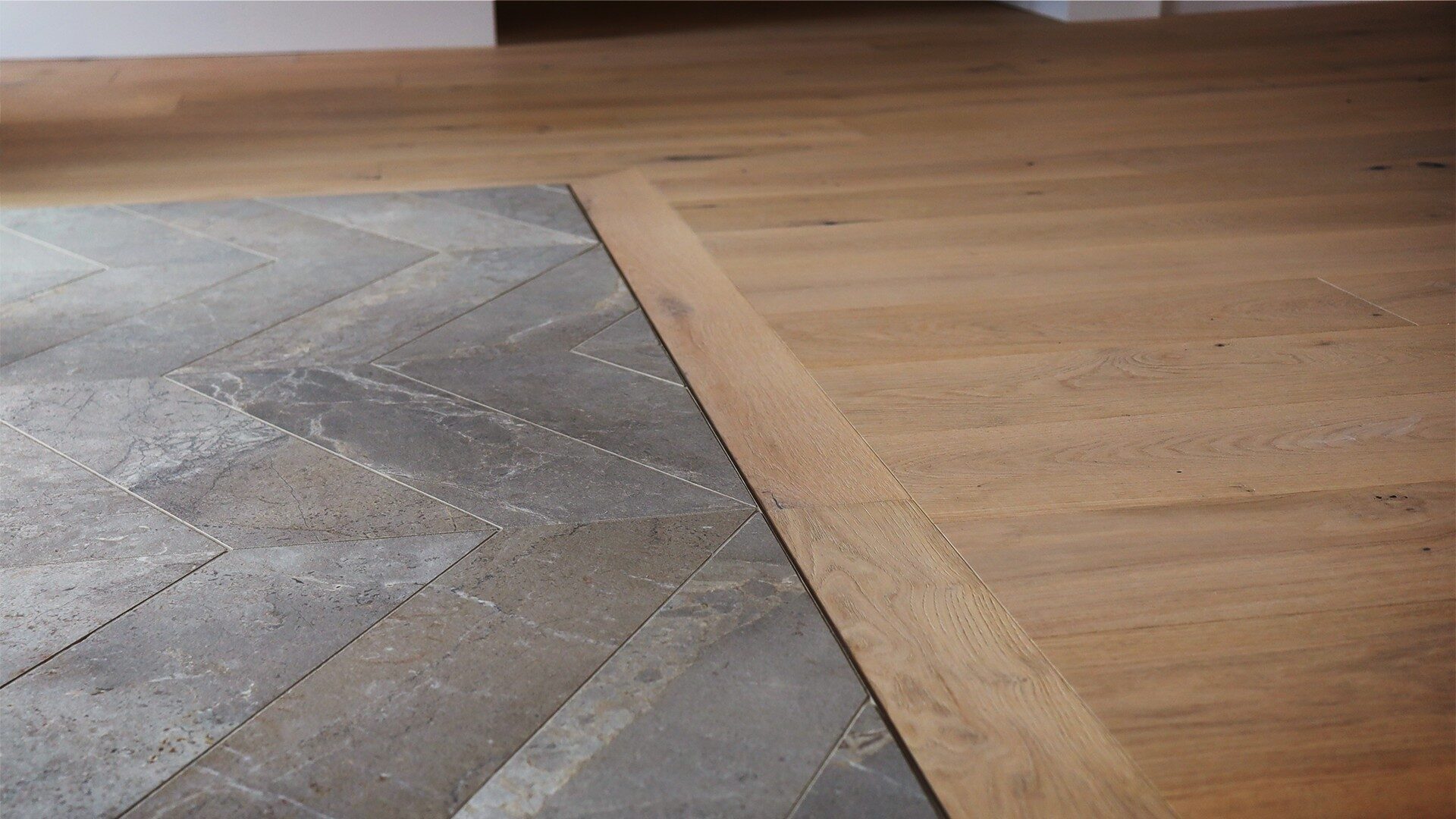 Close-up of a flooring transition between herringbone-pattern stone tiles and natural oak hardwood in an open-concept space.